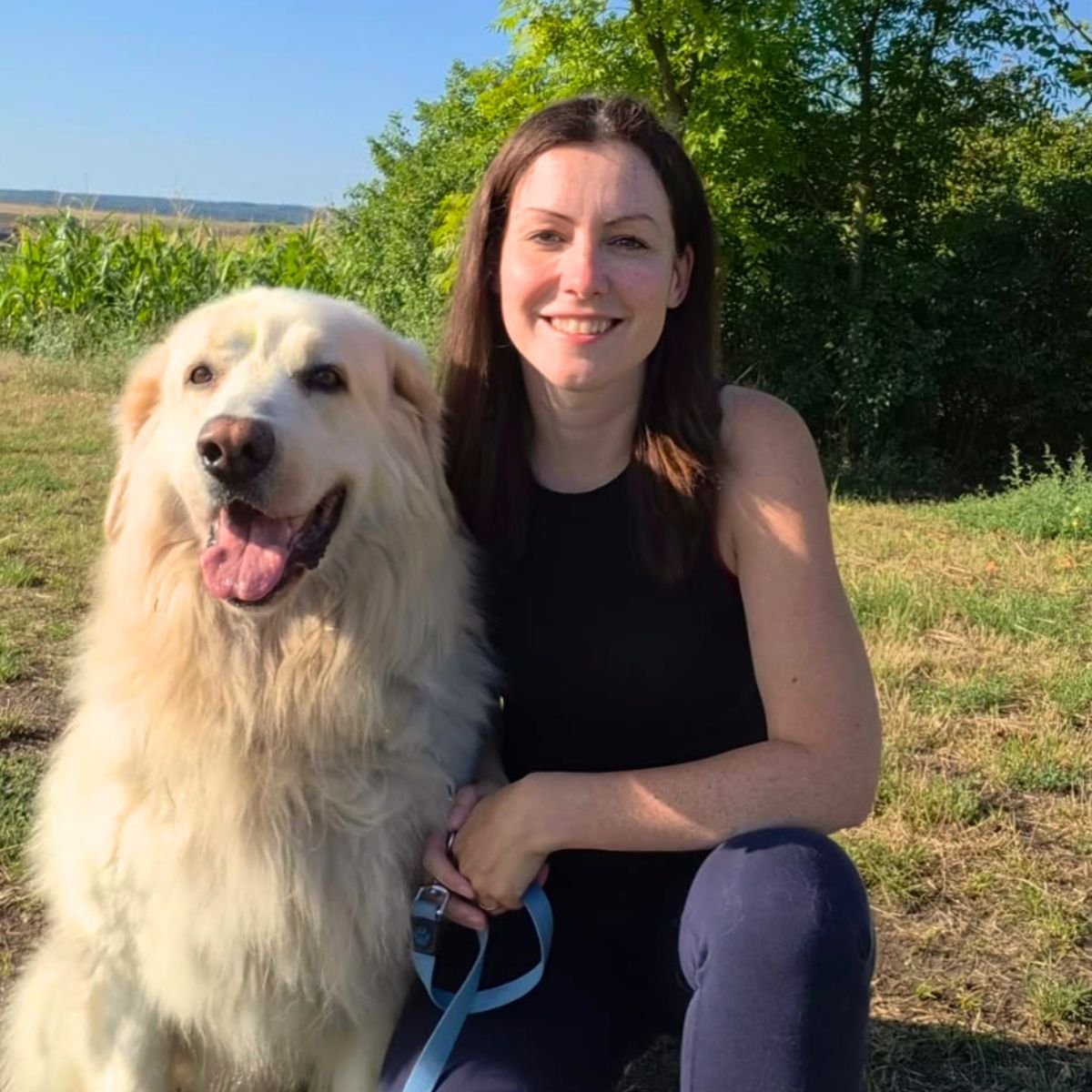 Zuza, founder of Gold Standard Dog Walking, kneeling outdoors beside a large white fluffy dog on a blue leash